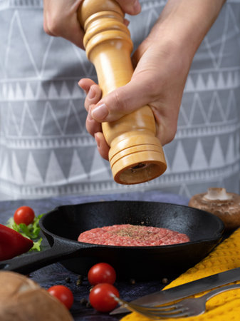 female hands use a wooden mill for spices, pray pepper on a meat patty raw in a cast iron panの写真素材