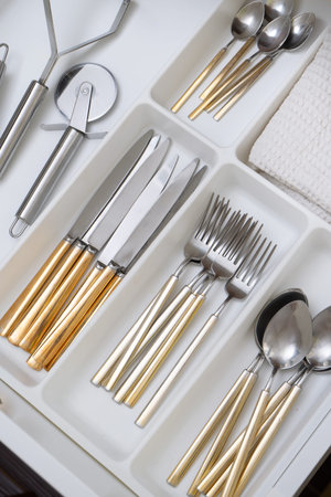 clean dishes in the drawer. Home cutlery set in gold and silver color, open home storage with crockery. Flat lay closeup with metal home furniture, spoon, fork and knife, and other kitchen gadgetsの写真素材