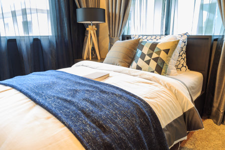 Many pillow and a book with white and brown blanket on the bed with brown wooden lamp side bed on...
