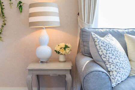 A modern lamp and flowers in a pot on white wooden table side a light blue fabric sofa with white patterned pillows and light through from blinds.の写真素材