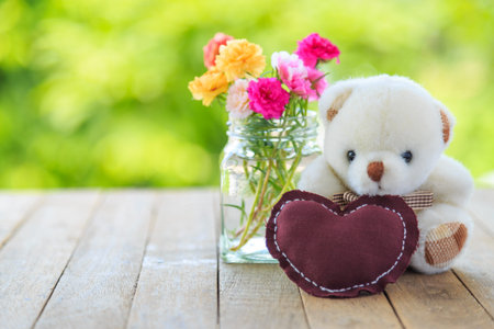 A white bear doll and brown heart pillow with moss rose flowers in glass vase on wooden table with...