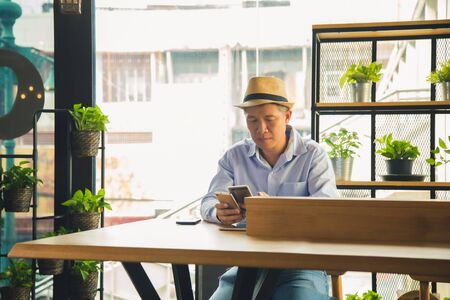 Medium close up portrait middle age Asian man in casual shirt is holding two mobile phone and confused from technology on wooden table at coffee shop with city building backgroundの写真素材