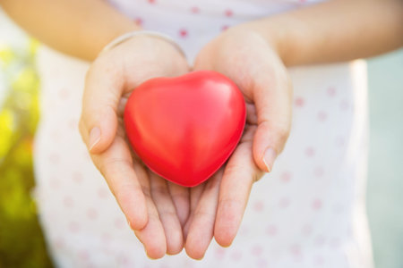 Close up a red heart shape ball in woman hands in a white pattern white dress blurred background.