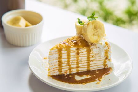 Close up side views a banana banoffee decorate by peppermint with sliced bananas in a white bowl and syrup bottle at the side on a white table next to the window with a nature background in the afternoon.の写真素材
