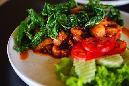 Close up Stir fried pork cracking and curry paste oin a white plate with tomato and cucumber topped with crispy basil decorated on a black table, Thai food conceptの写真素材