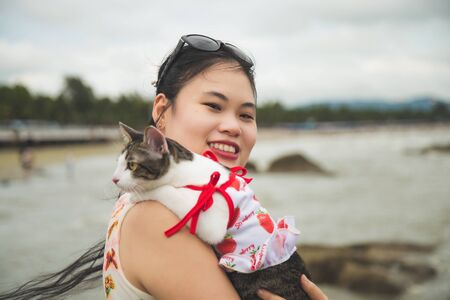 Close up portrait Smiley young Asian woman in a sweet dress standing and holding an adorable cat on her shoulder on the beach in the moody day.の写真素材