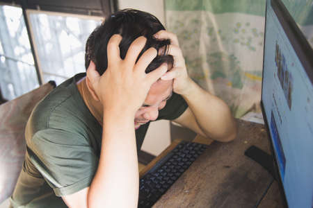 Close up high portrait extremely shocking Asian man in a green t-shirt sitting in front of the monitor computer on a wooden table at home.の写真素材
