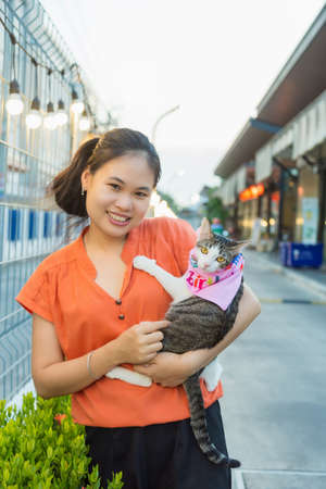 Close up portrait a long hair young Asian woman in orange shirt holding an adorable cat next to a metal fence on the way to parking at a food market in the evening.の写真素材