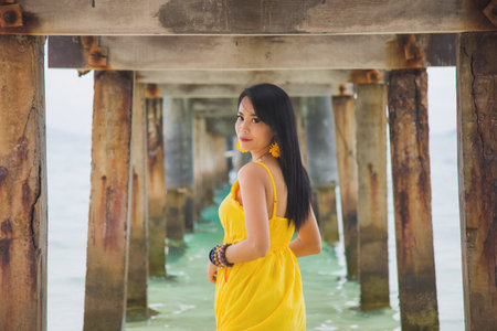 Portrait beautiful Asian woman in a yellow dress standing under the bridge over the sea, summer and...