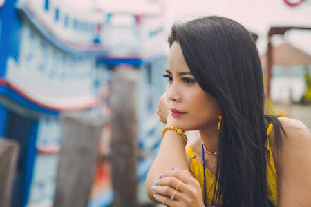 Close up Asian woman in a yellow dress put her chin on her hand looking away at a bay with sea boat blurred background.の写真素材