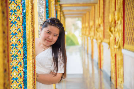 Smiling Asian woman in a white dress hides behind pillar in buddhist templeの写真素材