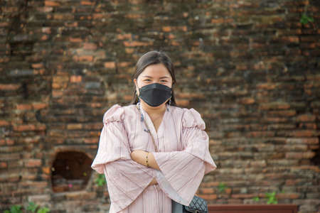 Asian woman in a pink dress cross her arms wearing a face mask with an old brick wall background.の写真素材
