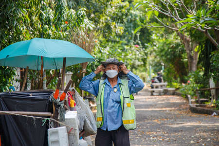 Portrait happy old Asian man street cleaner wearing a mask standing with his arms crossed next to an old gabage cart before going to work with nature background.の写真素材