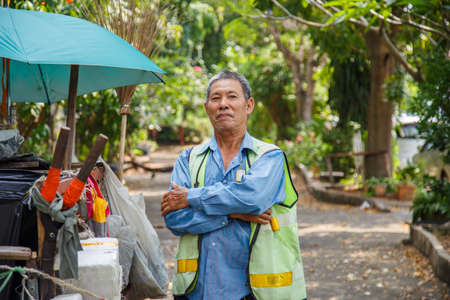 Portrait happy old Asian man street cleaner wearing a masks pick a pocket next to an old garbage cart before going to work with nature background.の写真素材