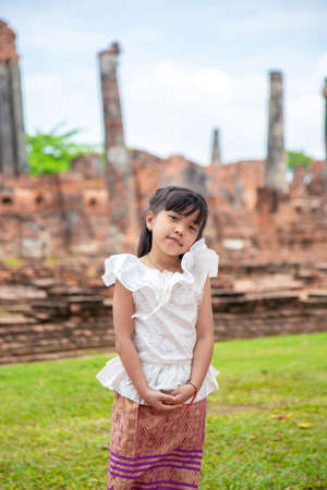 Portrait of Asian little girl in a white Thai shirt standing in front of an old pagoda while traveling in Asiaの写真素材