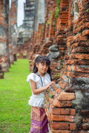 Portrait of Asian little girl in a white Thai shirt standing in front of an old pagoda while traveling in Asiaの写真素材