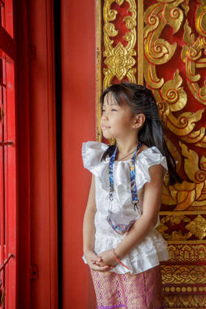 Portrait Asian little girl in a white Thai dress looking at side next to a beautiful wooden gate while traveling in Buddhist templeの写真素材