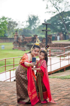 An Asian woman in a red Thai traditional dress take a picture with her daughter by her phone while traveling in Ayutthayaの写真素材