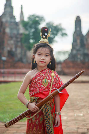 An Asian little girl in a red Thai traditional dress holding a Thai sword with ancient ruin backgroundの写真素材