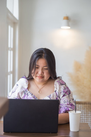 An Asian woman in a white patterned dress looking at outside window while working with laptop on a...