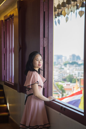 Portrait picture an Asian woman in a pink dress, she is looking at a camera  standing next to a window in temple while traveling in Asia.の写真素材
