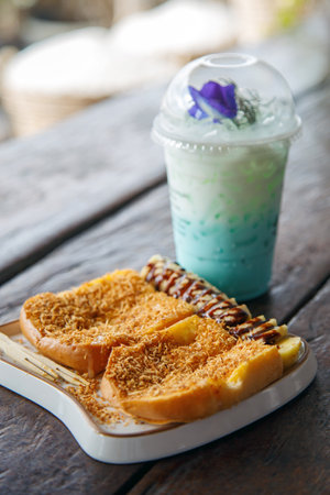 Toasted bread topped with roasted coconut and banana slices served with a glass of butterfly pea scented milk on a wooden table.の写真素材
