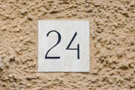 The number 24 is prominently featured on a square plaque attached to a rough-textured beige wall. Sunlight enhances the clarity of the details, showcasing its placement.の写真素材