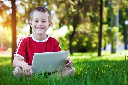 Cute happy six-year old boy lying with a laptop on the green grassの写真素材