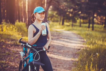 Young happy woman on bike standing on a hill road enjoying beautiful view summer sunny day resting drinking waterの写真素材