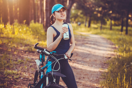 Young happy woman on bike standing on a hill road enjoying beautiful view summer sunny day resting drinking waterの写真素材