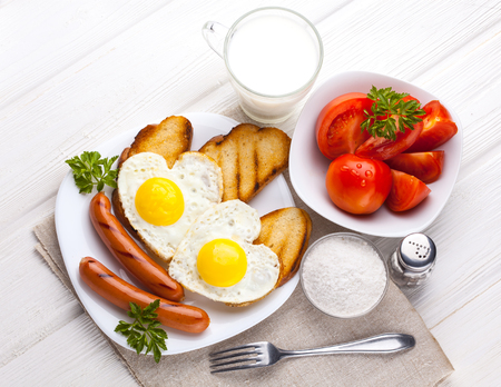 Breakfast on Valentine's Day - fried eggs in the shape of a heart and sausages, milk, tomatoes on the white wooden table.の写真素材