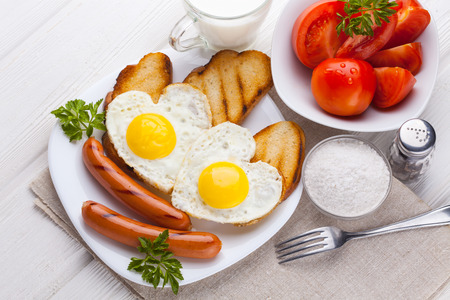 Breakfast on Valentine's Day - fried eggs in the shape of a heart and sausages, milk, tomatoes on the white wooden table.の写真素材