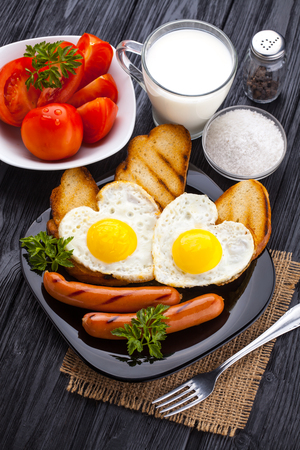 Breakfast on Valentine's Day - fried eggs in the shape of a heart and sausages, milk, tomatoes on the black wooden table.の写真素材