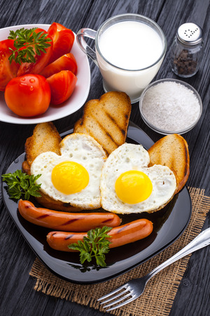 Breakfast on Valentine's Day - fried eggs in the shape of a heart and sausages, milk, tomatoes on the black wooden table.の写真素材