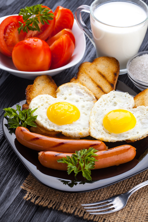 Breakfast on Valentine's Day - fried eggs in the shape of a heart and sausages, milk, tomatoes on the black wooden table.の写真素材
