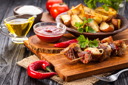 Meat skewer with herbs With onions, baked potatoes, tomatoes and greens on the black wooden tableの写真素材