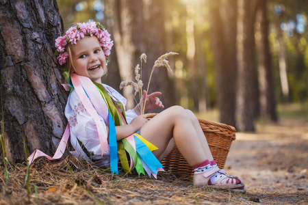 A little girl in a wreath with ribbons is sitting under a tree in the summer forest.の写真素材