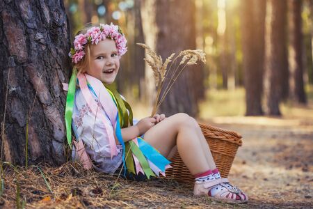 A little girl in a wreath with ribbons is sitting under a tree in the summer forest.の写真素材