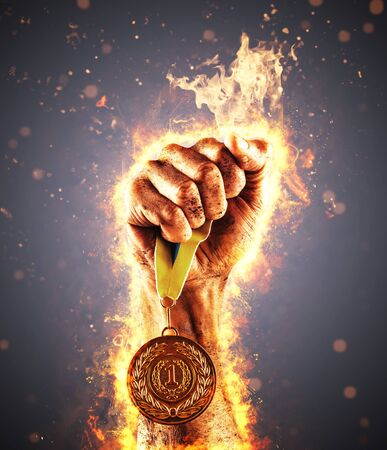 Man's hand in a fire is holding up gold medal on a dark background. Winner in a competition.の写真素材