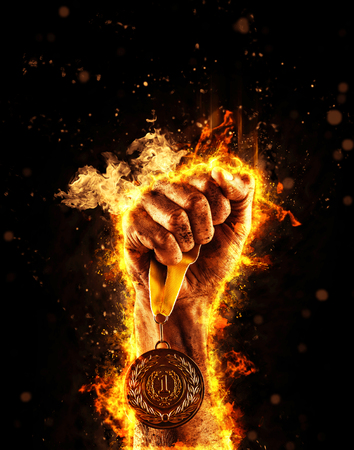 Man's hand in a fire is holding up gold medal on a dark background. Winner in a competition.の写真素材