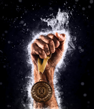 Man's hand in a blue fire is holding up gold medal on a dark background. Winner in a competition.の写真素材