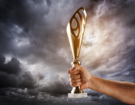 Mans hand is holding gold cup against of the gloomy stormy sky.の写真素材