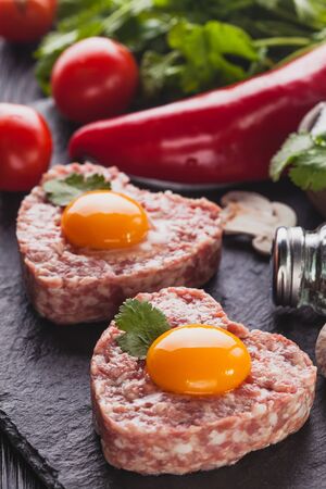 raw heart burger with egg on a black background with vegetables and herbs. Cookingの写真素材