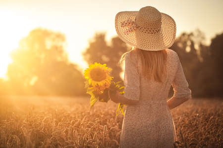 caucasian woman with sunflowers in a retro dress and a hat in a summer wheat fieldの写真素材