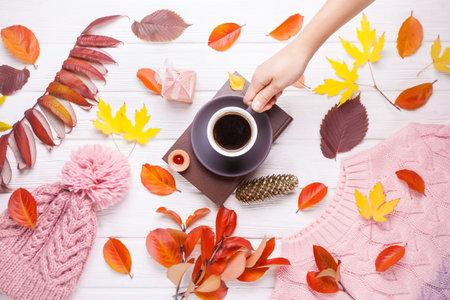 Book, a cup of coffee and a warm knitted hat, on an autumn wooden white table