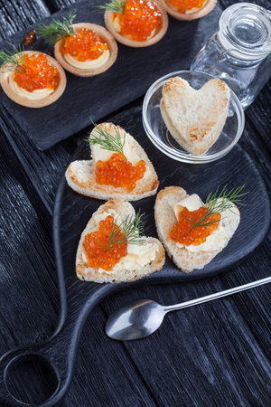 heart-shaped snacks with butter and red caviar served with dill on a black wooden tableの写真素材
