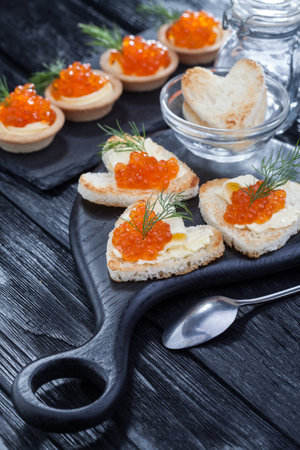 appetizer, heart-shaped snacks with butter and red caviar served with dill on a black wooden tableの写真素材