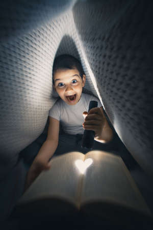 Little boy laying on the bed under blanket holding flashlight and reading book late at nightの写真素材