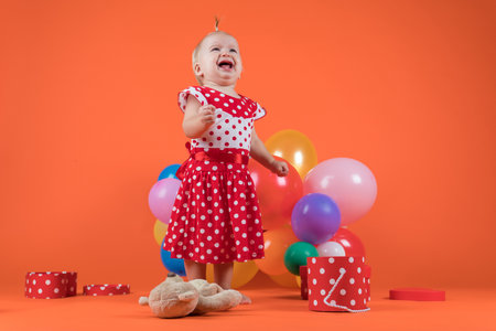 Crying baby girl in red dress upset with birthday gifts. Studio photo on orange backgroundの写真素材