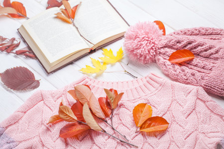 Book and a warm knitted hat and sweater, on an autumn wooden white table. Autumn background with yellow leavesの写真素材